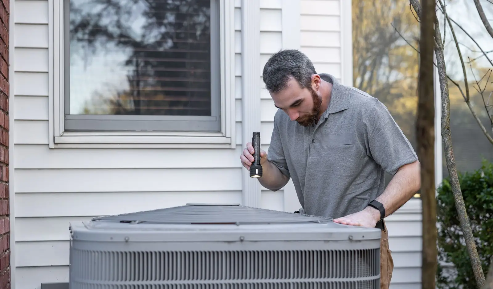 HVAC technician checking an outdoor condenser unit beside a house with a flashlight